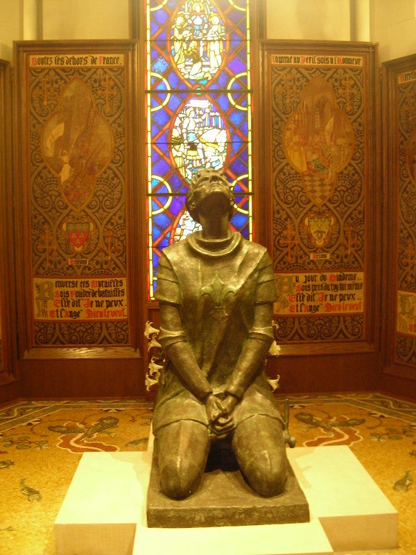 Statue of a kneeling Joan of Arc in front of stained glass panel