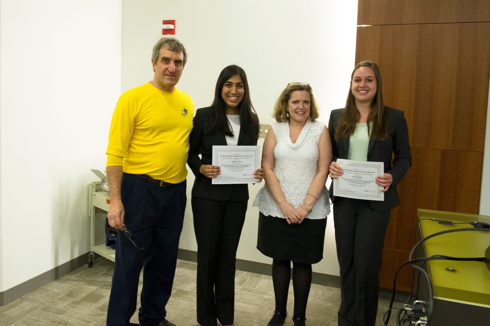 Budny, tall white man, graying hair, yellow shirt; Newborg, shorter white woman, blond hair in white shirt and dark skirt; with two award-winning, female-presenting students.