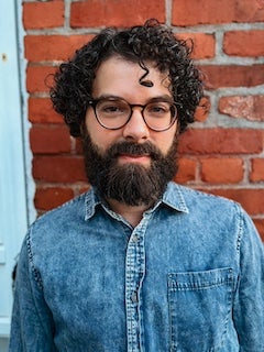 Adam Cetra is lightskinned with dark brown curly hair and beard/mustache, wearing dark-framed glasses, a light blue denim shirt and standing against a red brick wall.