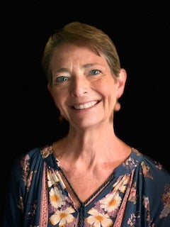 Phyllis Sigal is a white woman, with cropped brown hair, pink pendant earrings, and a bright smile against a black background.