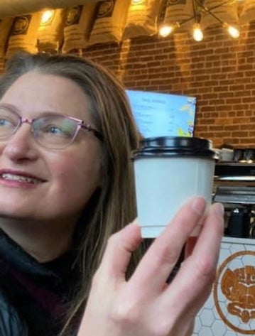 Ali Patterson, a white woman with pink eyeglass frames, holds a cup of takeout coffee