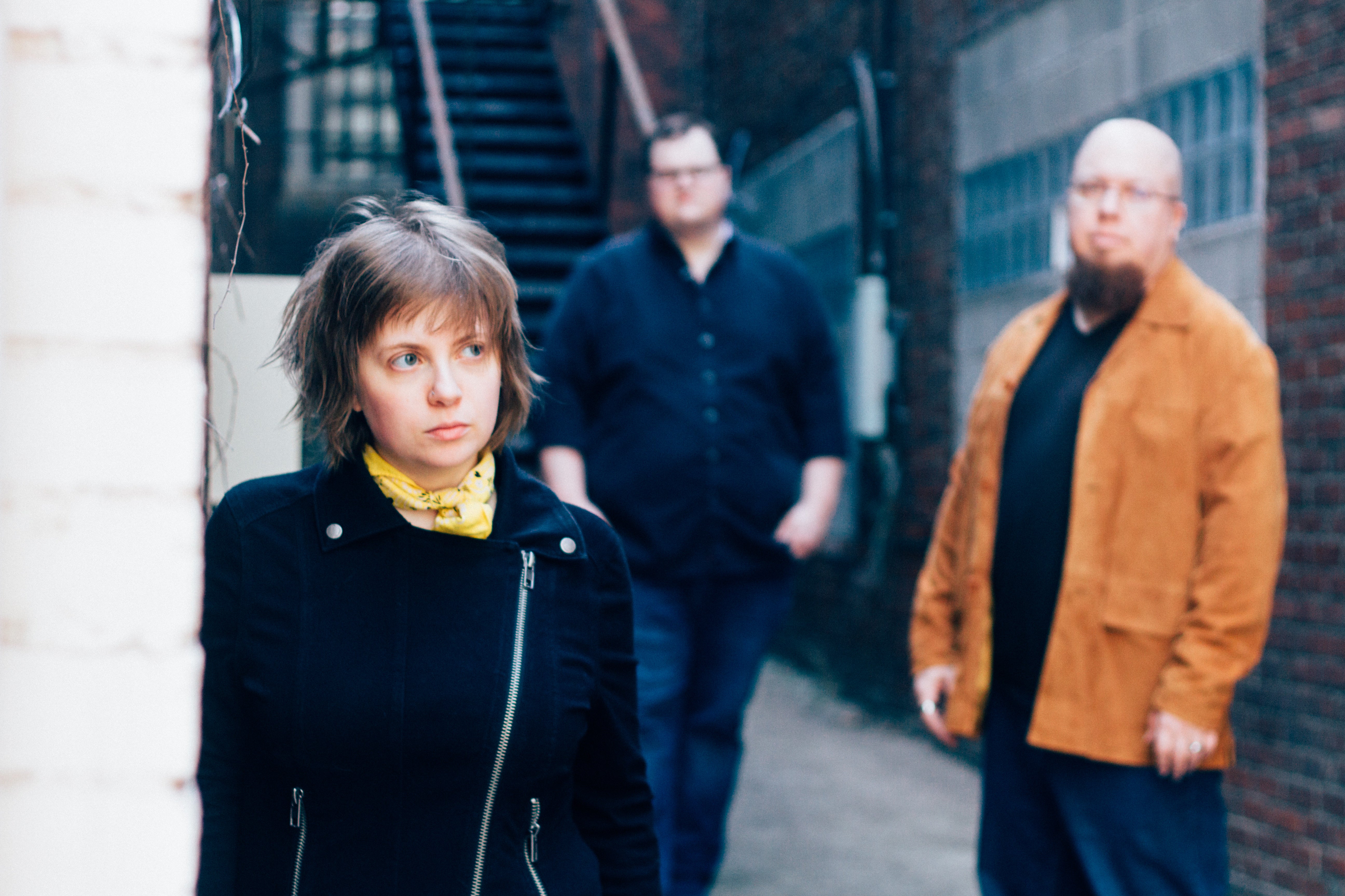 Lindsay Dragan on left, with shag-cut light brown hair, staggered to the right a band member and in background another band member; photo taken outside of a brick building with fire escape stairs 