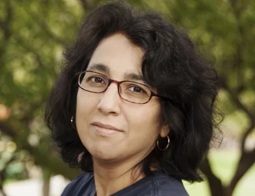 Geeta Kothari, a brown women with dark brown shoulder-length hair, auburn glasses, standing outdoors with trees in background