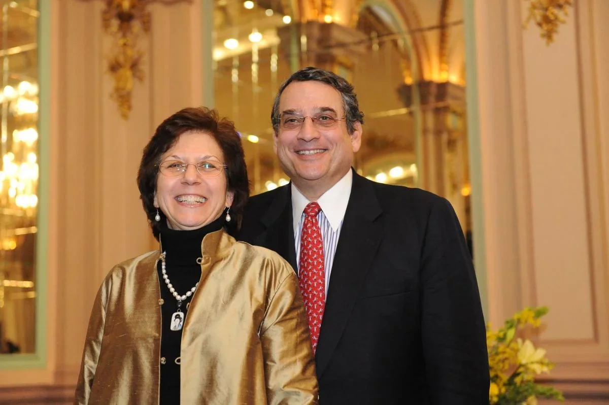 Tina Bellet with David Bellet: middle-Aged White Woman with short dark brown hair and glasses, wearing a black turtleneck under a gold jacket; middle-ged white man with salt and paper hair and glasses, wearing a dark suit with red patterned tie. 